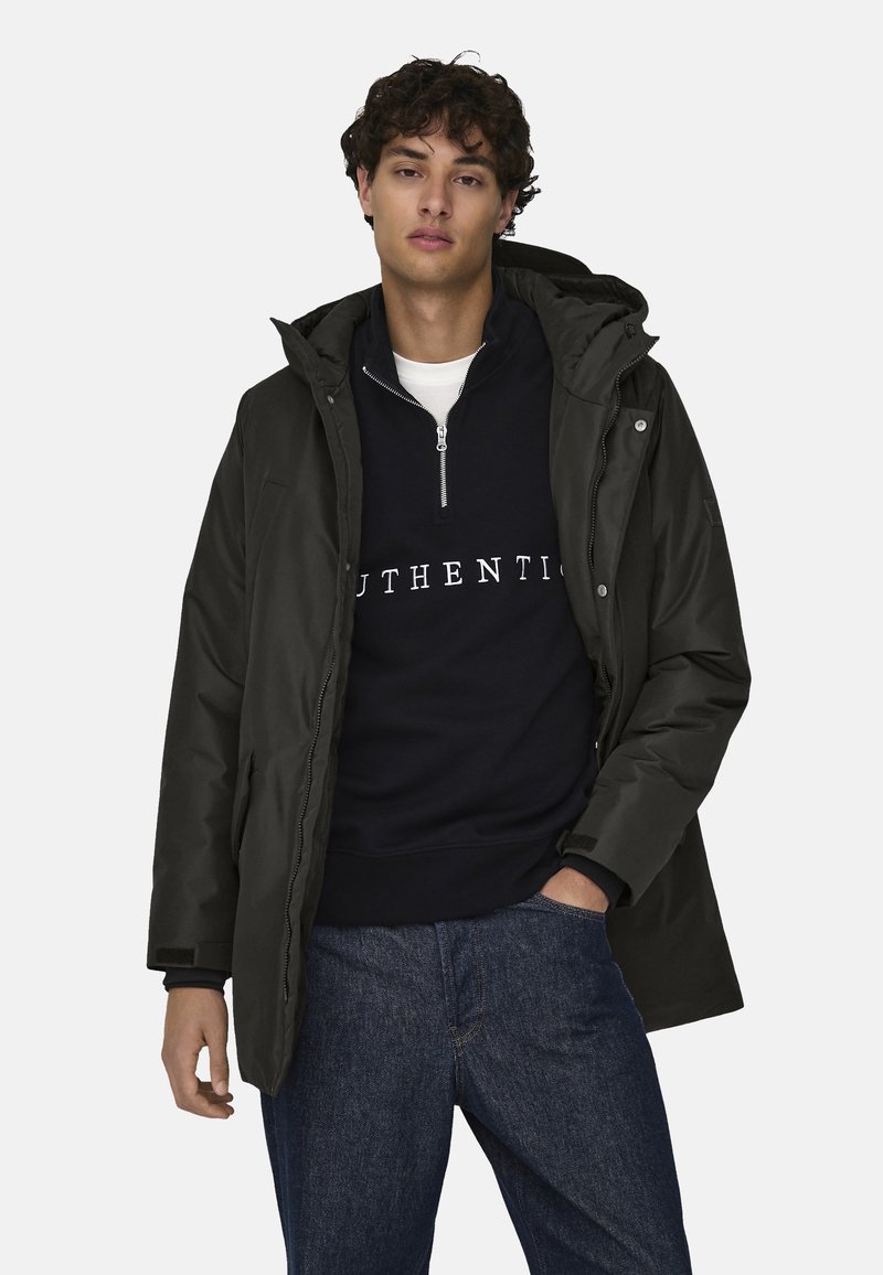 Sort parka med hætte, udstyret med lynlås foran og sidelommer, lagt over en mørk marineblå sweatshirt med hvid tekst "AUTHENTIC".