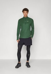 Grön långärmad sporttröja med dragkedja och grå sidopaneler, kombinerad med svarta shorts och kompressionsleggings. Svarta sportskor ingår.