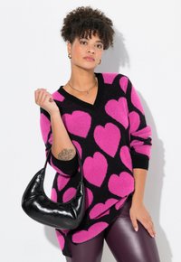Schwarzer und rosa herzgemusterter Pullover mit V-Ausschnitt und langen Ärmeln, kombiniert mit leuchtend lila Leggings und einer kleinen schwarzen Handtasche.