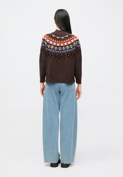 Inimene pikkade tumedate juustega, kes kannab pruuni mustrilist kampsunit ja laiade säärega hele siniseid teksapükse, seisab valge taustaga selg pööratuna.