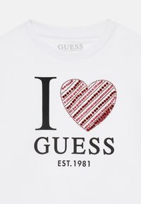 Valge puuvillane T-särk, millel on musta tekstiga "I love" ja punase sädelev südame kujundus, allpool on musta tekstiga print "GUESS EST. 1981".