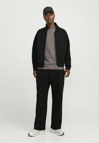 Veste zippée noire par-dessus une chemise grise, associée à un pantalon noir et des chaussures de sport argentées. Le modèle porte une casquette noire. Design simple, coupe décontractée.