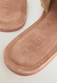 Sandalias de ante marrón con una textura suave, mostrando una puntera redondeada y costuras pronunciadas a lo largo del borde. Nombre de la marca estampado visible en la plantilla.
