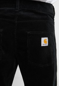 Pantalons en velours côtelé noir avec des côtes verticales, dotés de deux poches arrière et d'un patch logo jaune. Le tissu présente une apparence texturée.
