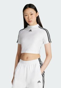 Biele skrátené športové tričko s čiernymi prúžkami na rukávoch, s vysokým stojacim golierom a logom adidas na prednej strane. Mäkko materiál, priliehavý strih.