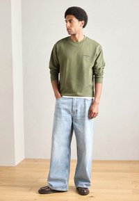 Sudadera verde oliva con cuello redondo y detalle bordado, combinada con vaqueros de pierna ancha azul claro y zapatos marrón oscuro.