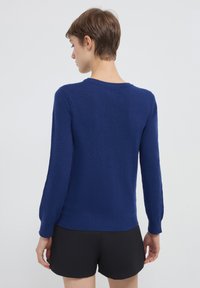 Person, der mit dem Rücken zur Kamera steht, trägt einen langärmeligen blauen Pullover und schwarze Shorts und steht vor einem schlichten hellen Hintergrund.