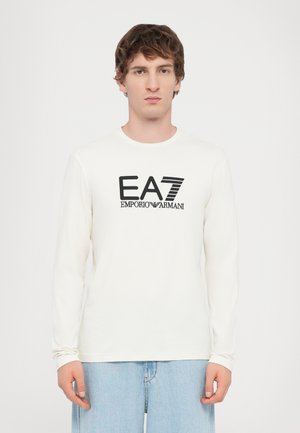 White long-sleeve shirt featuring a black EA7 Emporio Armani logo. Cotton material with a round neckline and relaxed fit.