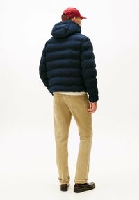 Chaqueta de plumas azul marino con capucha ajustada, que presenta amplias secciones acolchadas. Combinada con pantalones beige claros y calzado negro.