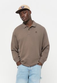 Brown long-sleeve polo shirt with a two-button placket and embroidered logo, paired with light blue denim shorts. Smooth texture.