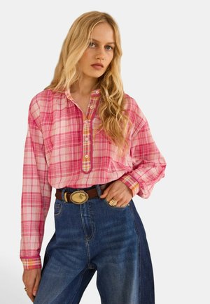 Donna bionda che indossa una camicia a quadri rosa a maniche lunghe infilata in jeans blu scuro a vita alta con cintura marrone e fibbia dorata.
