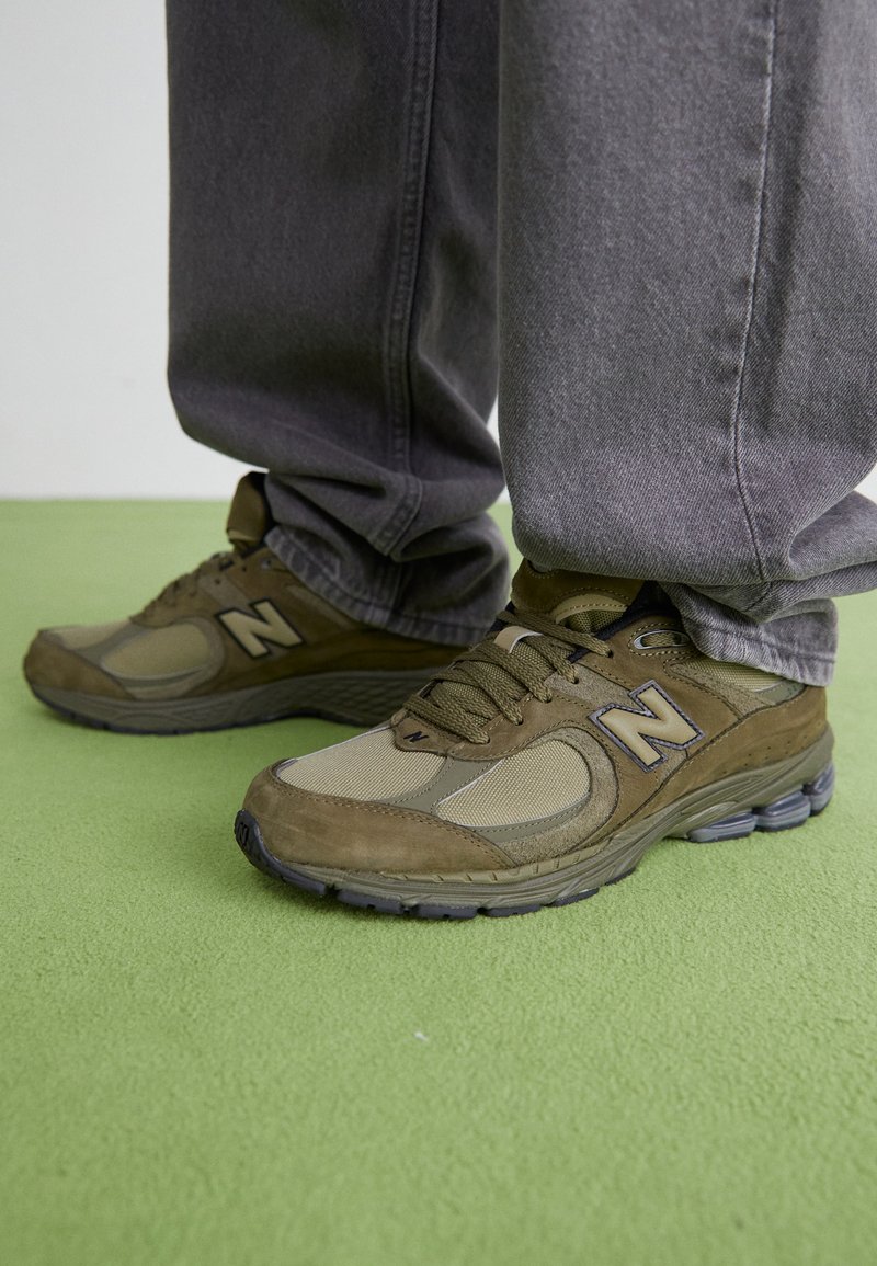 Des baskets New Balance vert olive présentent un mélange de matériaux en daim et en mesh, avec une semelle rembourrée et un logo 'N' bien visible sur le côté.