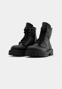 Czarne skórzane buty typu combat na sznurówki z grubymi kauczukowymi podeszwami i bocznymi zamkami, pokazane na jednolitym białym tle.