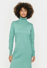 Gaiši zaļa adīta kleita ar augstu apkakli. Garās piedurknes, riboti aproces, pieguļošs siluets un maiga tekstūra.