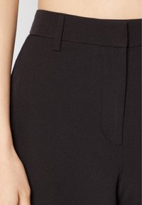 Närbild på svarta byxor med hög midja, bältesöglor och dold framknäppning, båda på ljus hudton.