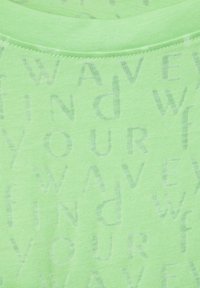 Světle zelené tričko s texturou a opakujícím se potiskem slov "FIND YOUR WAVE" v různých odstínech zelené. Hladká tkanina s kulatým výstřihem.