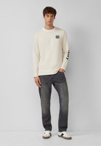 Lichtbeige longsleeve shirt met een zwart logo op de borst en tekst op de mouw, gecombineerd met donkergrijze jeans en witte sneakers.