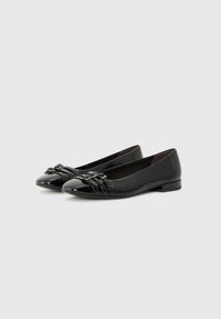 Ballerines en cuir noir avec un bout arrondi, des accents de nœud décoratifs et un talon plat. La surface a une finition brillante et des détails texturés.
