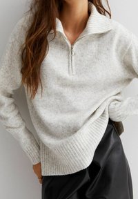Hellgrauer, gerippter Strickpullover mit einem Halbreißverschluss am Kragen. Verfügt über einen weichen, strukturierten Stoff und einen lässigen Schnitt, kombiniert mit einem schwarzen Rock.