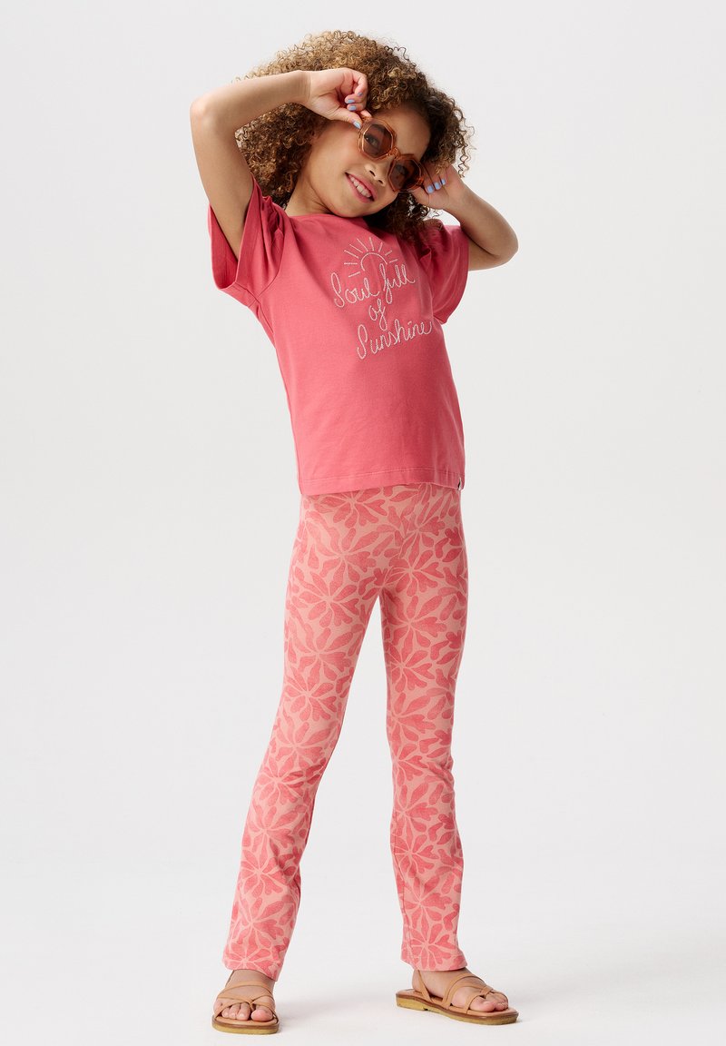 Roze T-shirt met een zonnenafbeelding, gecombineerd met roze leggings met een bloemenpatroon. Bruine sandalen maken de outfit compleet.