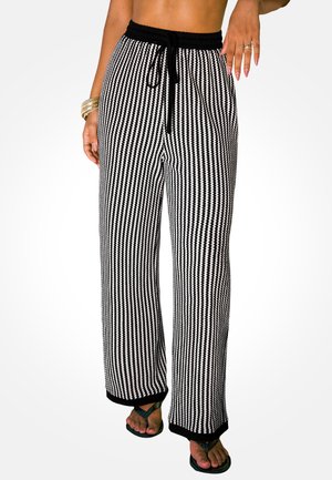 Femme portant un pantalon large à rayures verticales noires et blanches avec une taille à cordon noir et des sandales noires, montrant le ventre et les mains.
