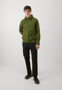 Groene fleecejas met capuchon, ritssluiting aan de voorkant en een logo op de borst. Gecombineerd met zwarte cargobroek met zijzakken. Zwarte schoenen.