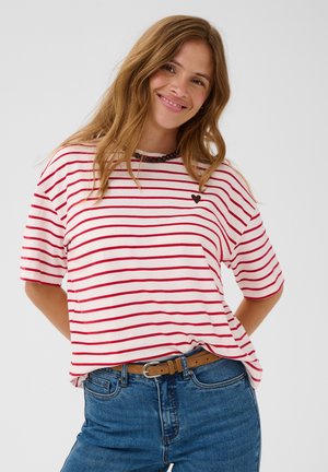 Femme souriante portant une chemise blanche à rayures horizontales rouges et un petit cœur noir, un jean bleu et une ceinture marron sur fond blanc.