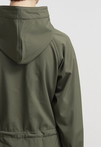 Personne portant une veste à capuche vert olive, vue de dos avec des coutures visibles et une taille froncée.