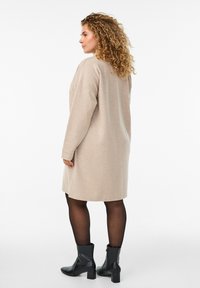 Žena s kovrčavom kosom odjevena u bež haljinu dugih rukava, crne prozračne čarape i crne čizme do gležnja, stoji na bijeloj pozadini.