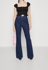 Černý květinový crop top s nabíranými rukávy, v kombinaci s vysokými rozšířenými modrými džíny. Modelka drží malou růžovou kabelku a má na sobě černé špičaté boty.