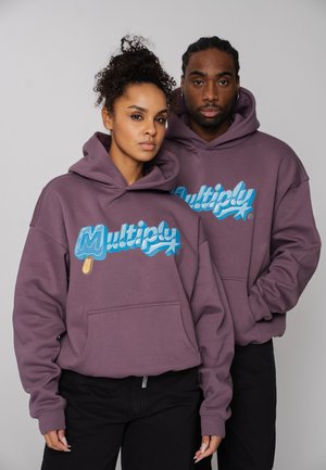 Two people wearing matching purple hoodies with a blue "Multiply" logo against a plain gray background, standing close together.