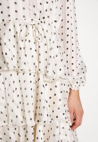 Blusa de capas en tejido semitransparente color crema con lunares negros. Presenta capas con volantes, mangas con puños y un cierre de botón con detalle de lazo.