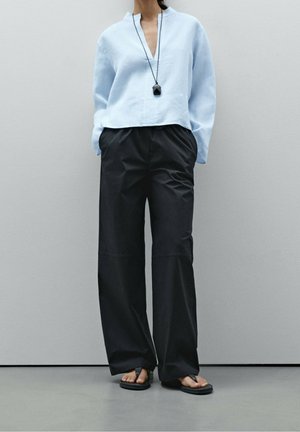 Person wearing a light blue long-sleeve blouse, black wide-leg pants, black sandals, and a long pendant necklace, standing against a plain wall.