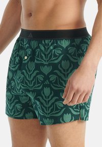Gröna boxershorts med ett blommönster i mörkare grönt, en svart elastisk midjeband och en funktionell sidoficka med en knapp.