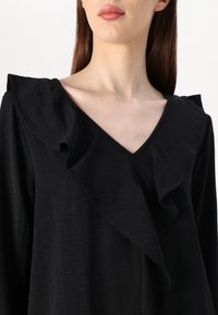Črna bluzica z V-izrezom, ki ima ruffle ovratnik, dolge rokave in teksturirano tkanino. Gladek in lahek material s subtilnim sijajem.