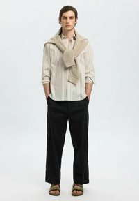 Camicia a righe in tonalità chiare, maglione beige appoggiato sulle spalle, pantaloni neri larghi e sandali verdi. Tessuti morbidi al tatto.