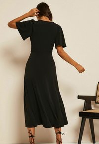 Musta midi-mekko, jossa on lyhyet, liehuvat hihat, pehmeä kangas ja väljä siluetti. Ominaisuuksiin kuuluu sileä tekstuuri ja hienovarainen liike.