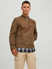 Giacca marrone in suede con colletto alto, zip frontale e tasche laterali. Indossata sopra una camicia a quadri, abbinata a pantaloni cargo beige.