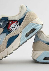 Sneakers mają beżową cholewkę z niebieskimi akcentami, texturedny materiał i pluszową naszywkę z postacią. W podeszwie znajduje się widoczna poduszka powietrzna.