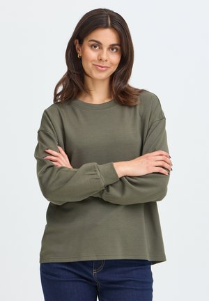 Ung kvinde med mørkt hår iført en grøn langærmet trøje og blå jeans, stående med armene krydsede, med et let smil, foran en ensfarvet baggrund.