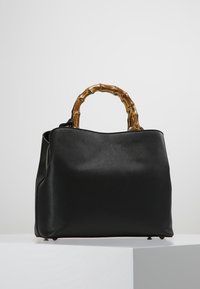 Černá texturovaná kabelka s zakřivenými rukojeťmi ve stylu bambusu, strukturovaným tvarem a zlatými nožičkami, umístěná na bílé podstavě proti šedému pozadí.