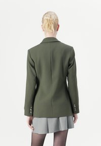 Olivgrön kavaj med en figurnära design, enfärgad fram och tre-knapplösning på ärmarna, tillsammans med en grå plisserad kjol.