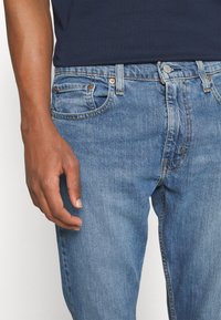 Main masculine reposant sur le côté, portant un jean bleu et une chemise bleu marine foncé, montrant la taille avant et le haut de la cuisse.