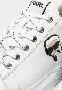 Λευκό αθλητικό παπούτσι από δέρμα με υφή στη σόλα που έχει ασημί στίγματα, επίπεδα κορδόνια, μεταλλικά ρούχνια και μικρό λογότυπο χαρακτήρα σε ασπρόμαυρο.