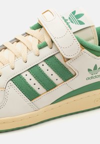 Sneaker bianca in pelle con dettagli in suede verde, tre strisce verdi, lacci beige e un cinturino in velcro. Presenta una suola testurizzata e un branding discreto.