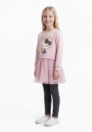 Fată tânără zâmbitoare cu păr lung și blond, purtând un hanorac roz Hello Kitty, fustă din tulle roz, colanți închiși la culoare și pantofi albi slip-on.