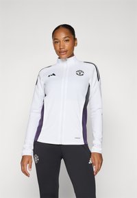 Veste zippée blanche en tissu léger, avec des panneaux latéraux noirs et violets, logo sur la poitrine et poignets côtelés. Associée à un pantalon sombre.