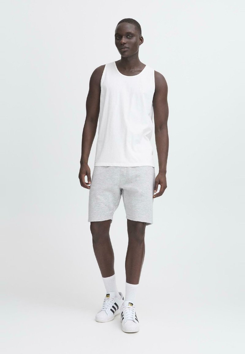 Hvit ermeløs tanktop, lysegrå shorts og hvite sneakers med svarte striper. Modellen står mot en lys bakgrunn.