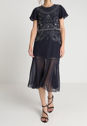 Marineblaue Midikleid mit filigraner silberner Stickerei, flatternden Ärmeln und transparentem Chiffonrock, kombiniert mit schwarzen, hochhackigen Riemchensandalen.