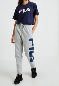 Kobieta ubrana w granatowy t-shirt FILA, jasnoszare dresy z dużym granatowym logo FILA oraz czarne sneakersy FILA, stojąca z jedną ręką w kieszeni.